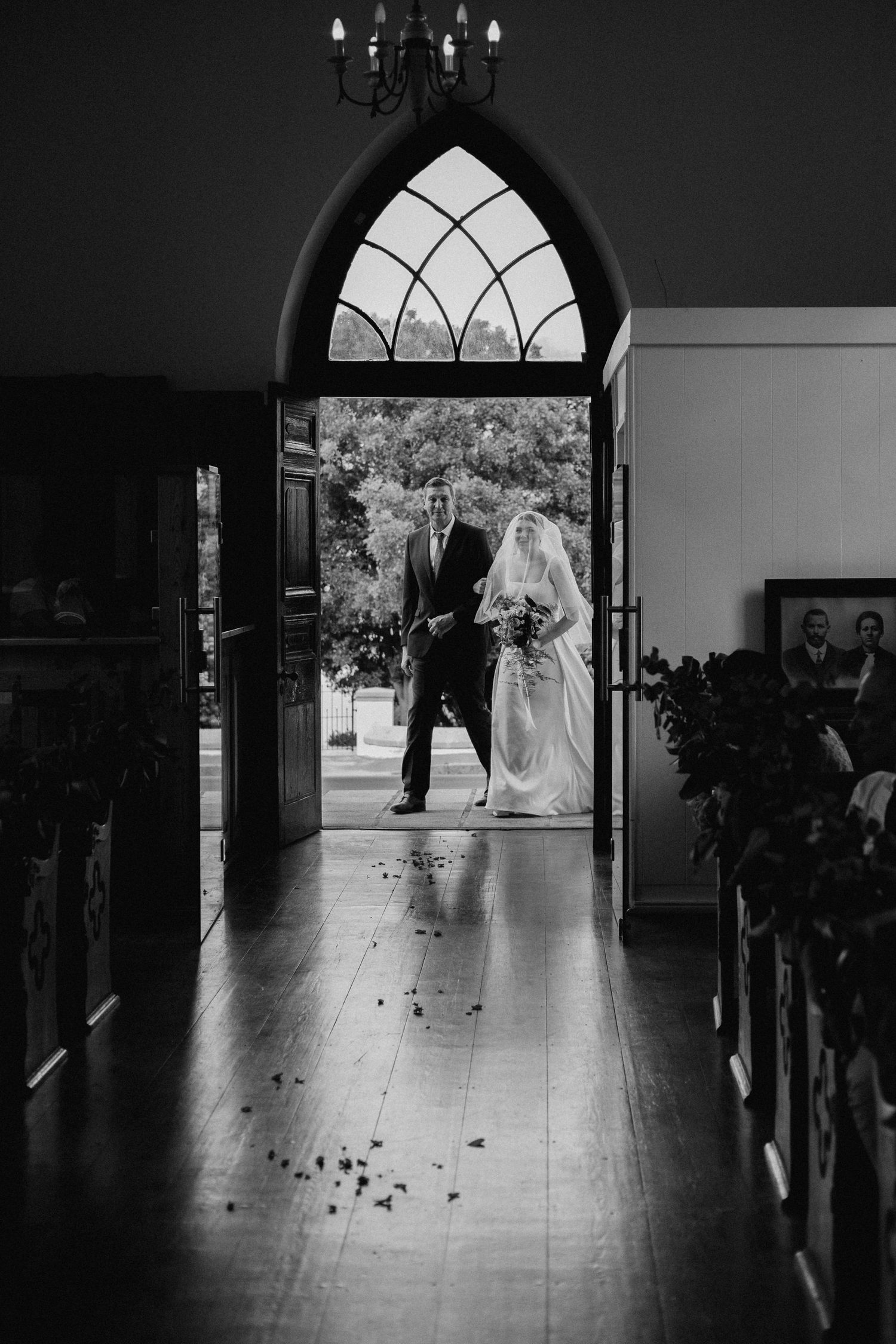 A black and white church interior with silhouettes standing in a gothic arched doorway on a flower-strewn aisle.