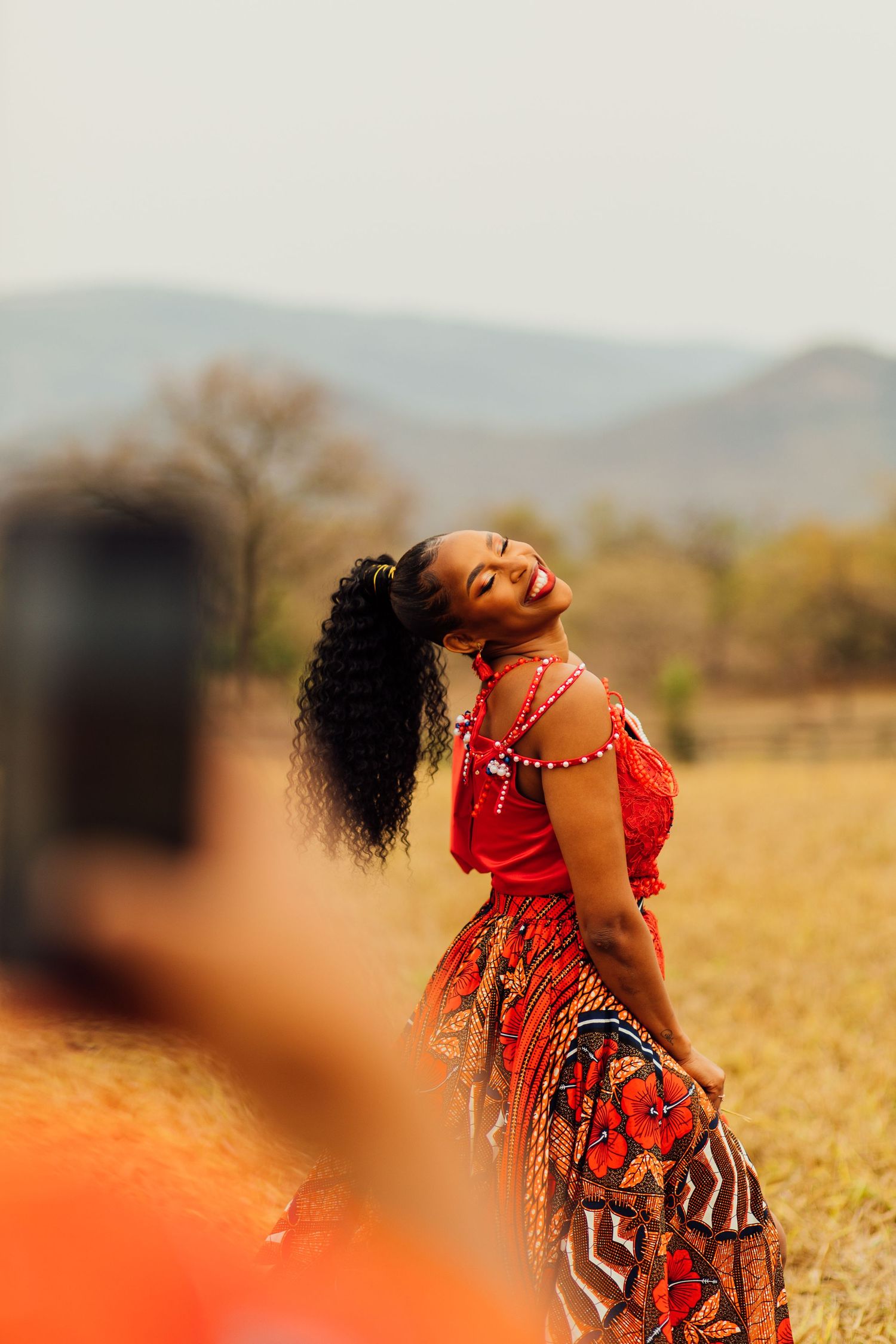 Woman in vibrant patterned dress smiles joyfully in golden field with mountains visible in background.