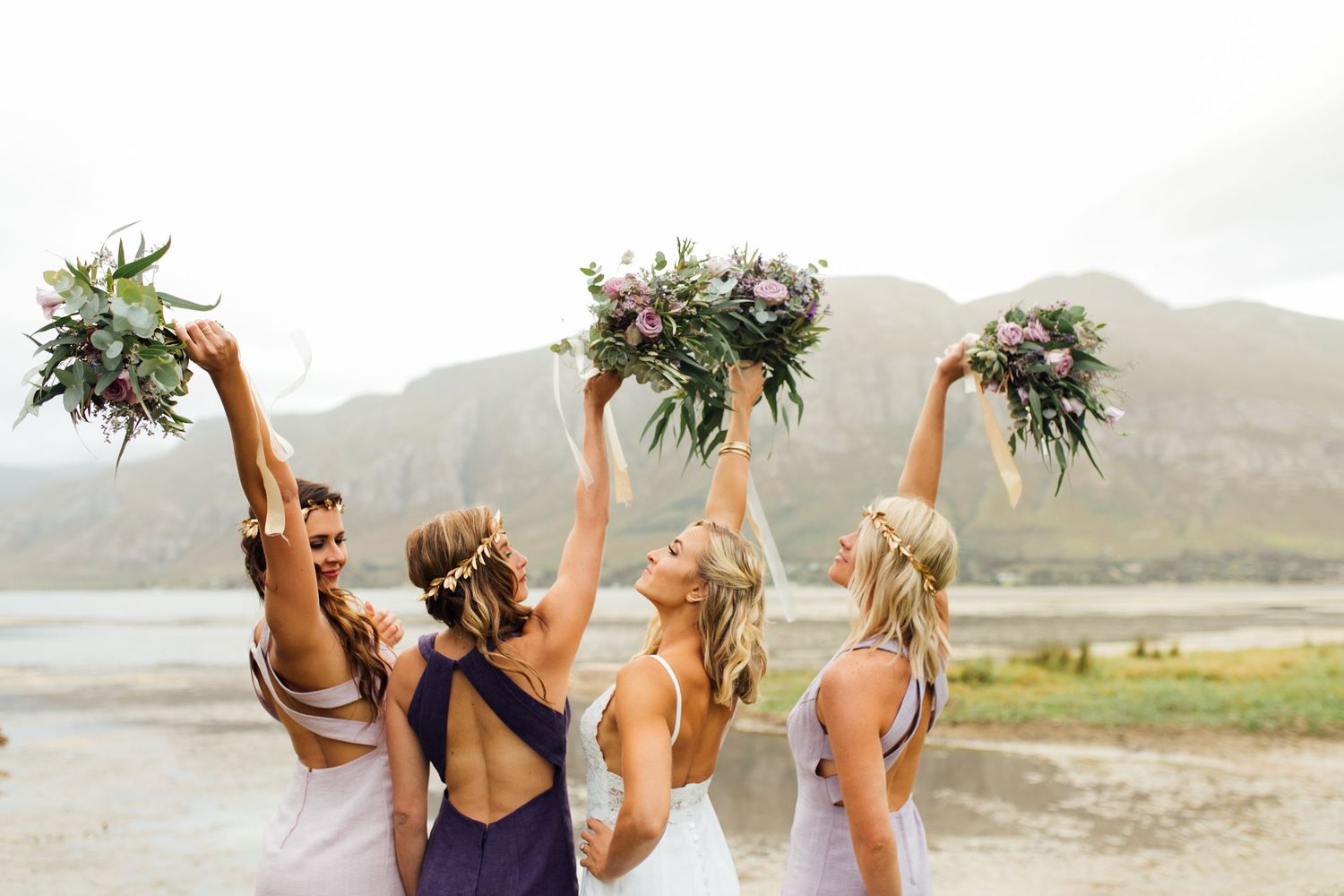 Three bridesmaids in purple dresses raise their flower bouquets against a misty mountain landscape.