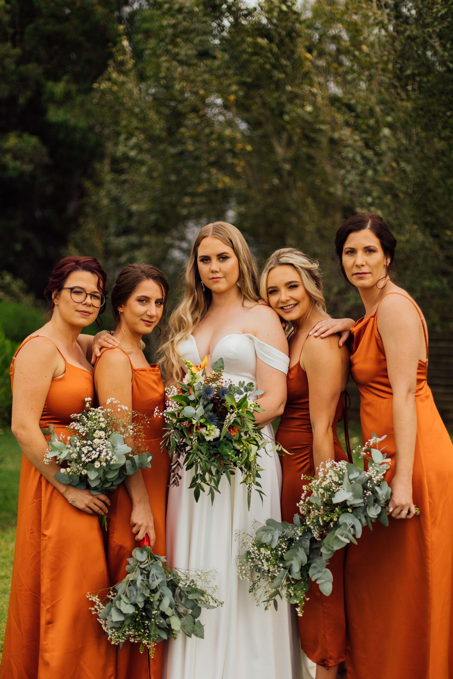 Bridal party wearing burnt orange bridesmaid dresses holding lush greenery bouquets with eucalyptus in an outdoor setting.