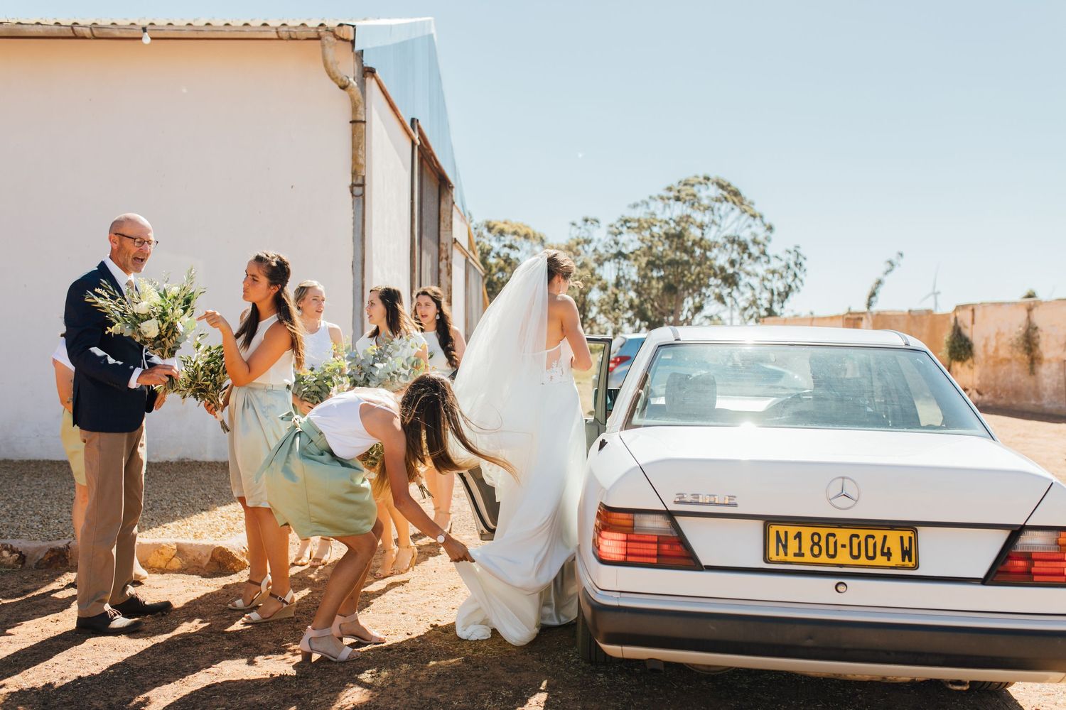 Wedding party gathers around a silver car on a sunny day outside a white building, with flowing dress in the breeze.