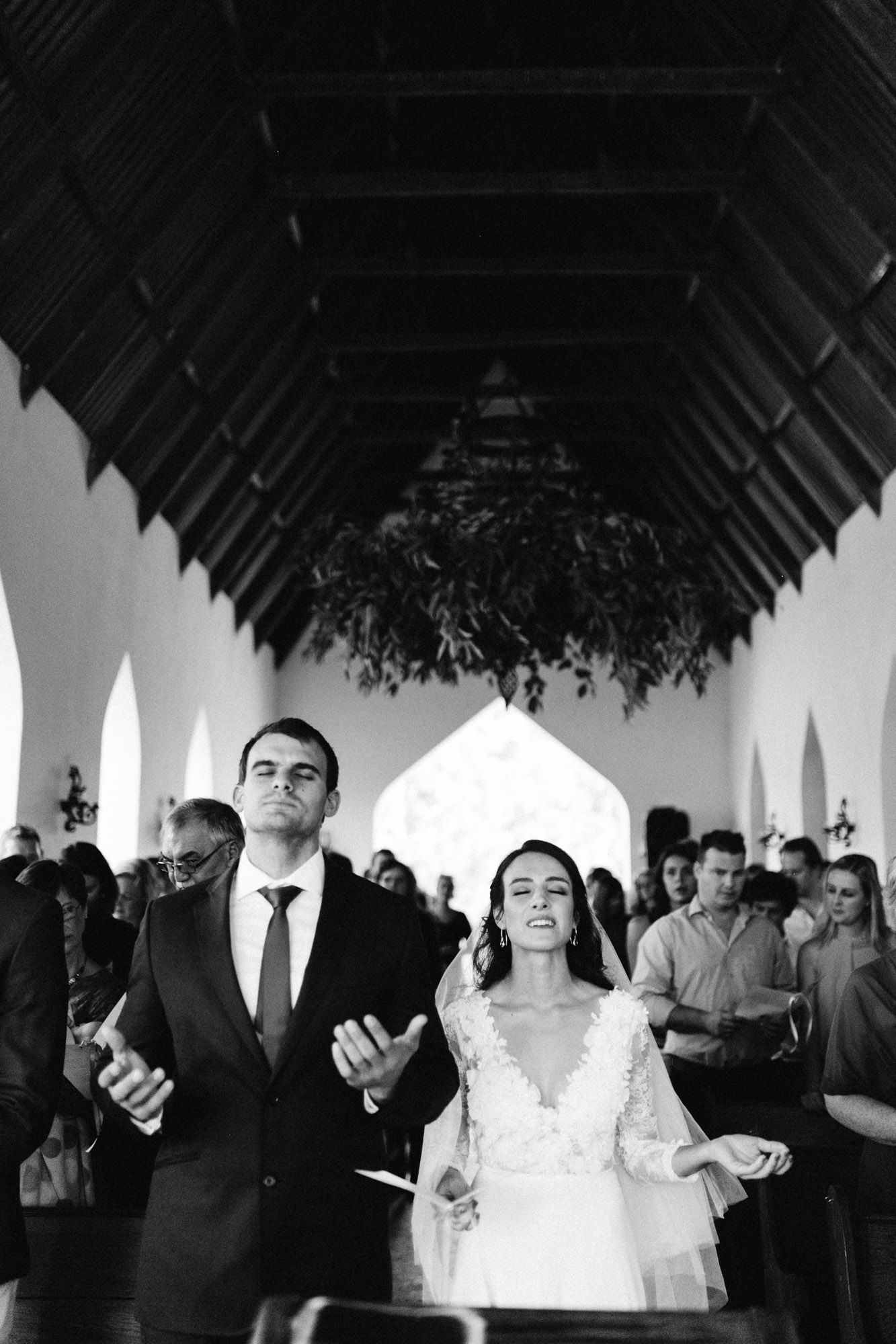 Inside a church with vaulted ceiling and arched windows, a couple prays together during their black and white wedding ceremony.