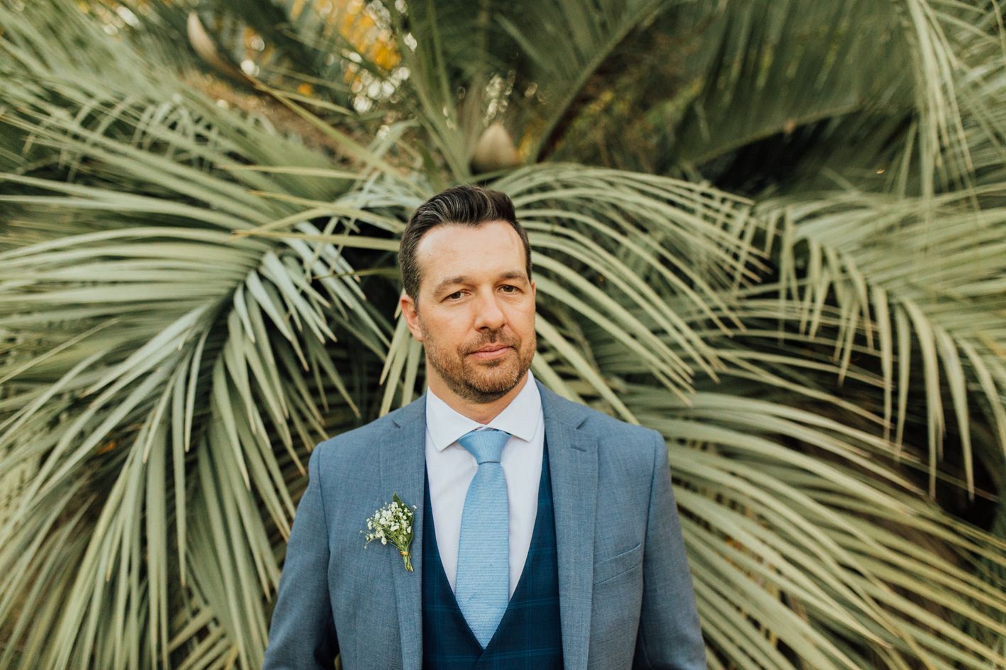 A groom in a blue suit and tie poses in front of tropical palm fronds during an outdoor wedding.