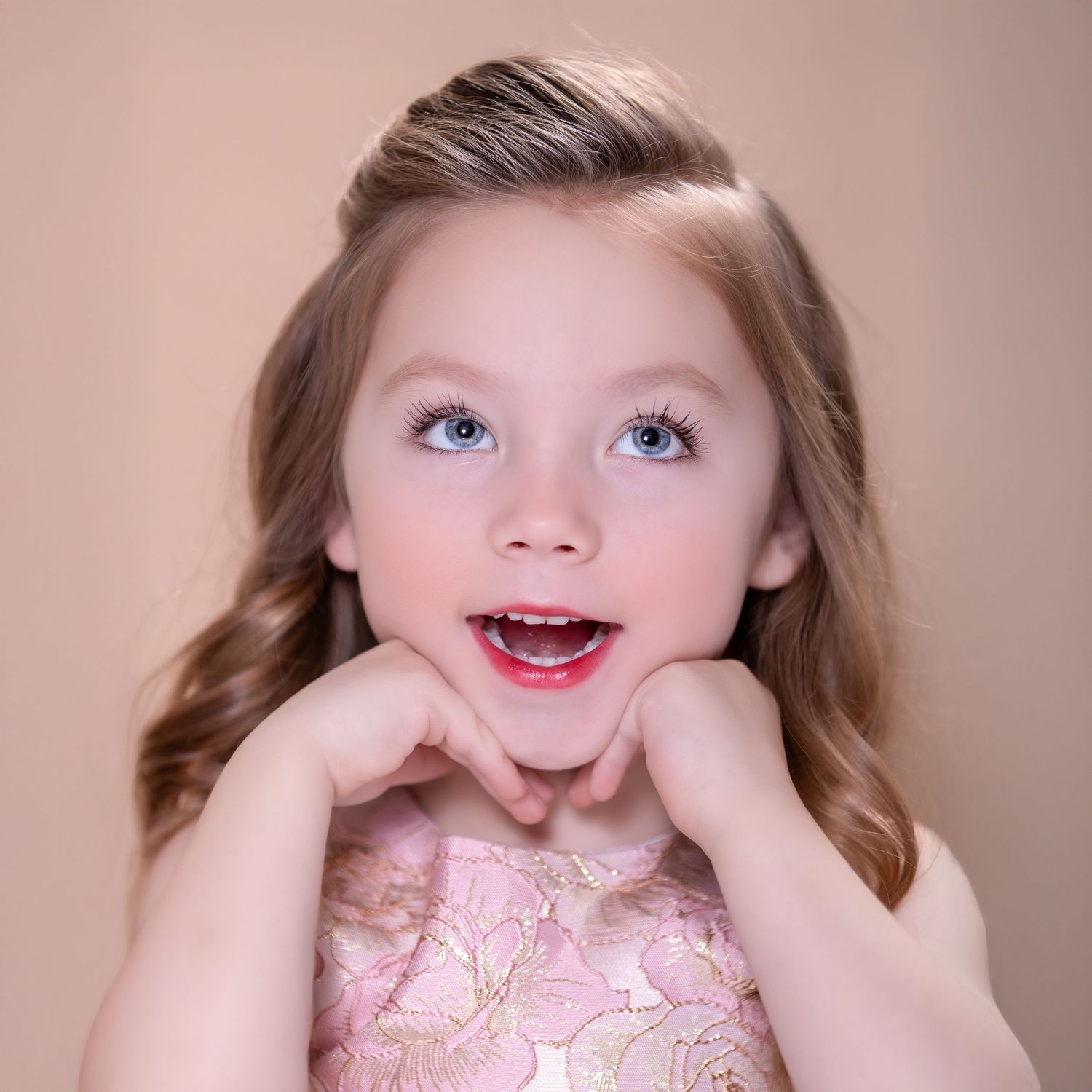 A young child brings hands to face in an expressive pose while wearing a pink lace dress against a neutral background.