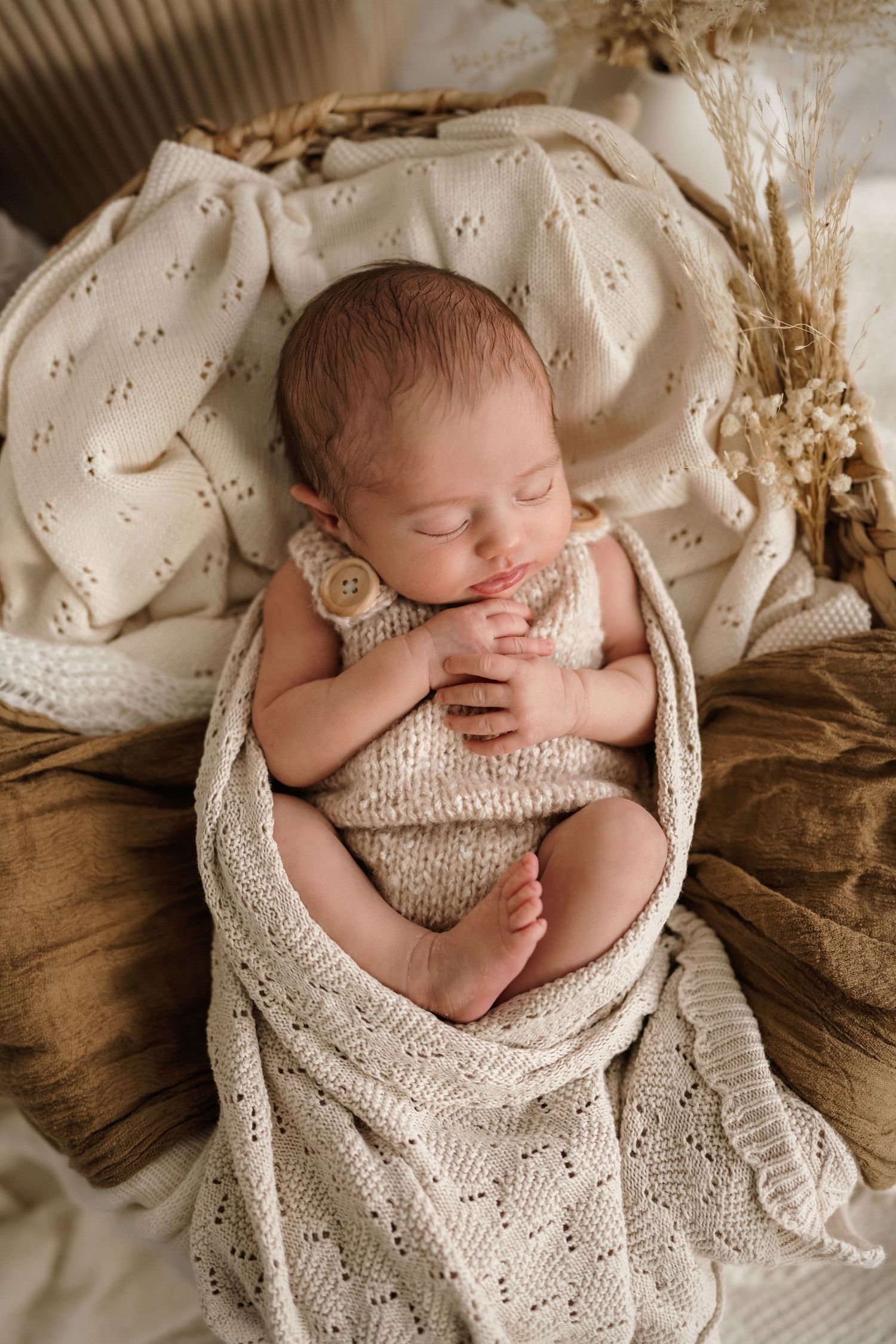 Sleeping newborn in white wrap nestled in knitted brown basket.