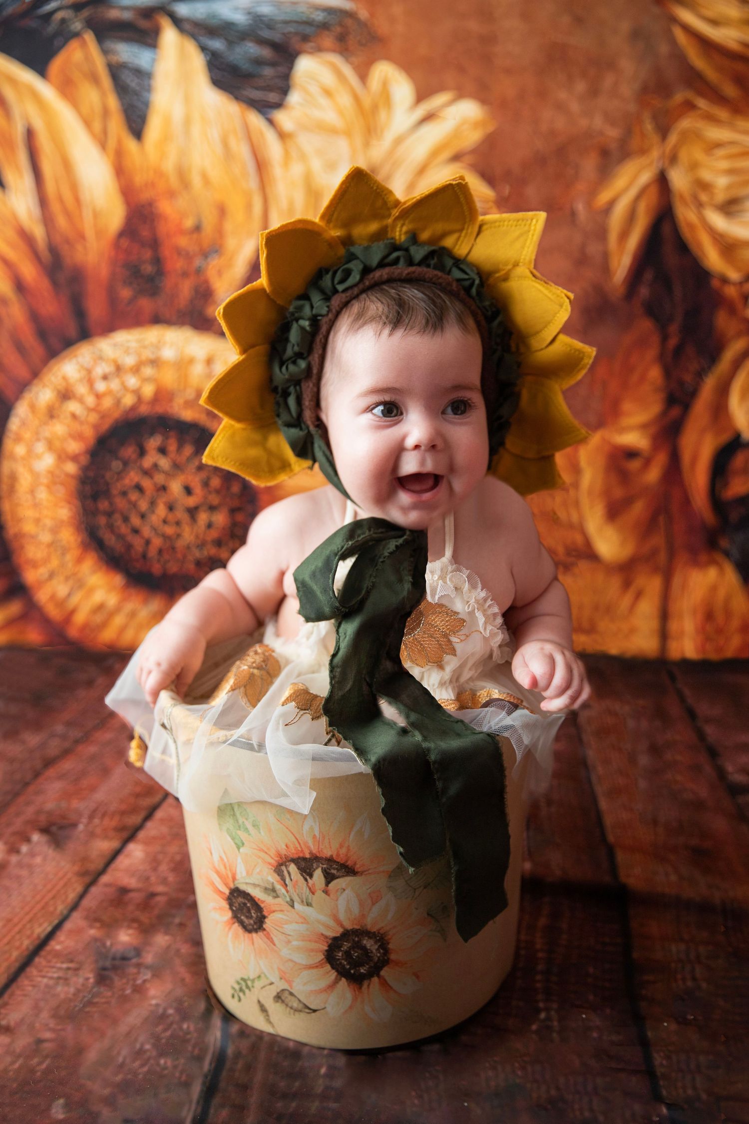 Adorable baby in sunflower bonnet sitting in decorative floral bucket with sunflower painted backdrop.