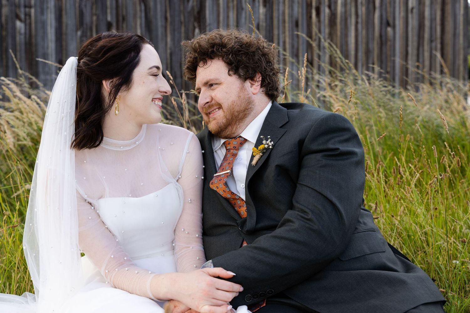A bride and groom sit together smiling outdoors, she in a white gown with veil, he in a dark suit with floral boutonniere.