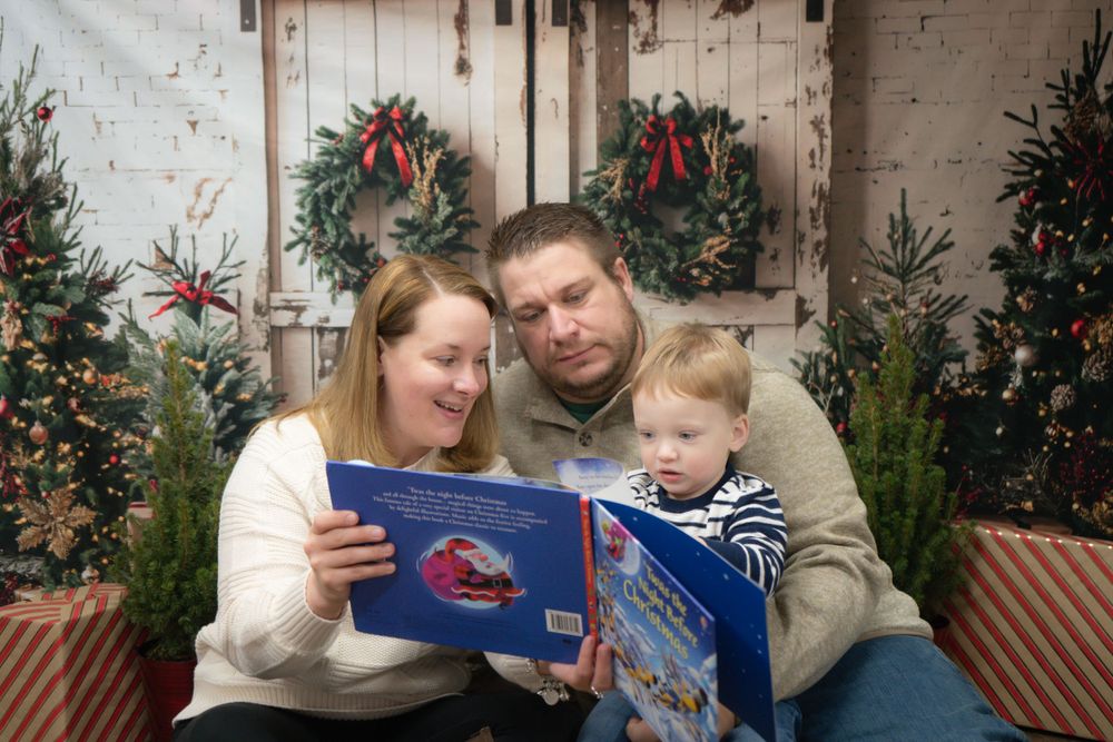 A family reads a Christmas story together in front of a decorated holiday scene with wreaths and evergreen trees.