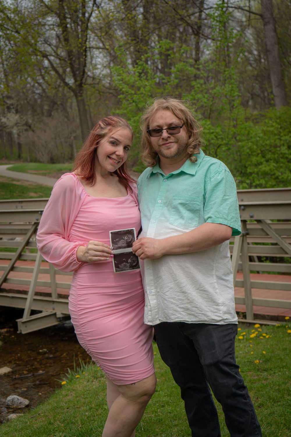 A couple stands on a wooden bridge in a park with spring foliage, sharing a tender moment together.