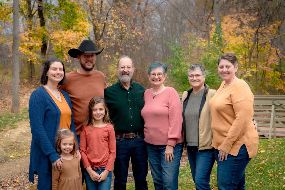 A group of people wearing colorful sweaters pose together outdoors on an autumn day with fall foliage in the background.