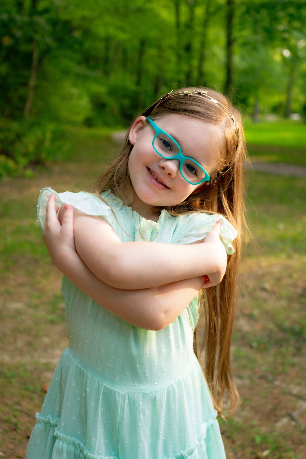 Young child in mint green dress and blue glasses posing playfully outdoors.