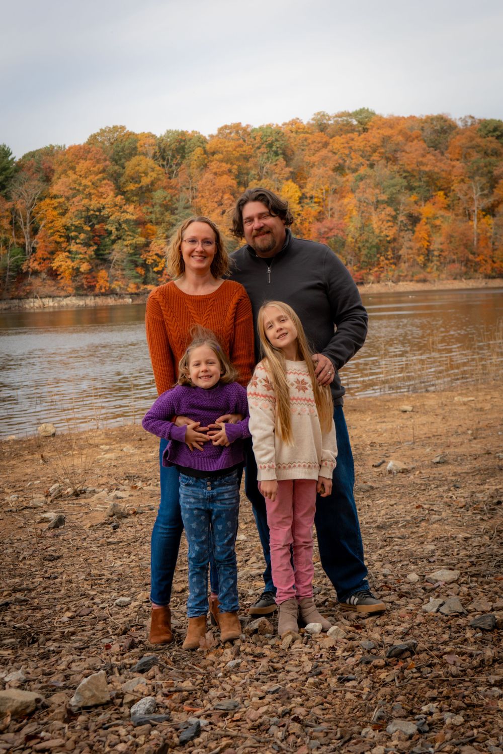 A family stands together on a rocky shoreline with vibrant fall foliage and a lake in the background.