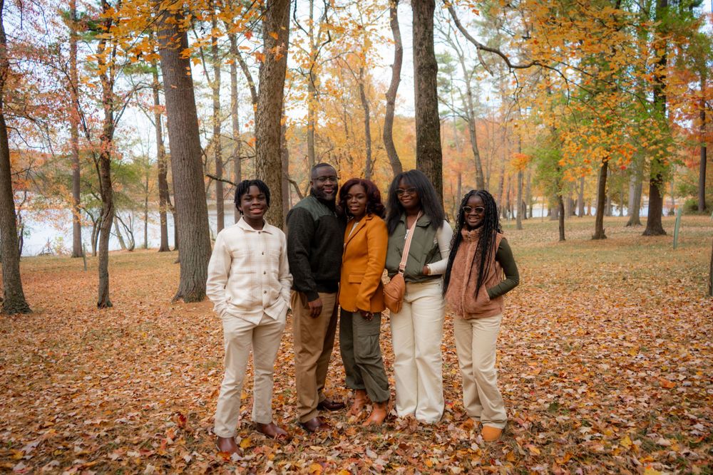 A group stands together on fallen autumn leaves in a park with orange foliage and trees surrounding them.