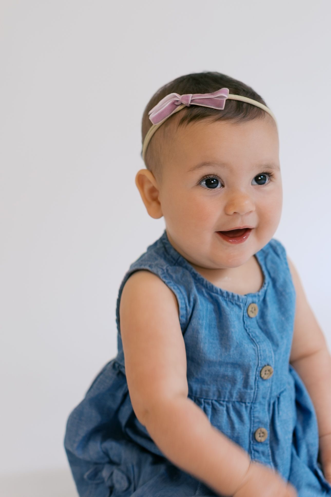 Smiling baby girl in blue denim dress and pink bow headband sitting against white background.