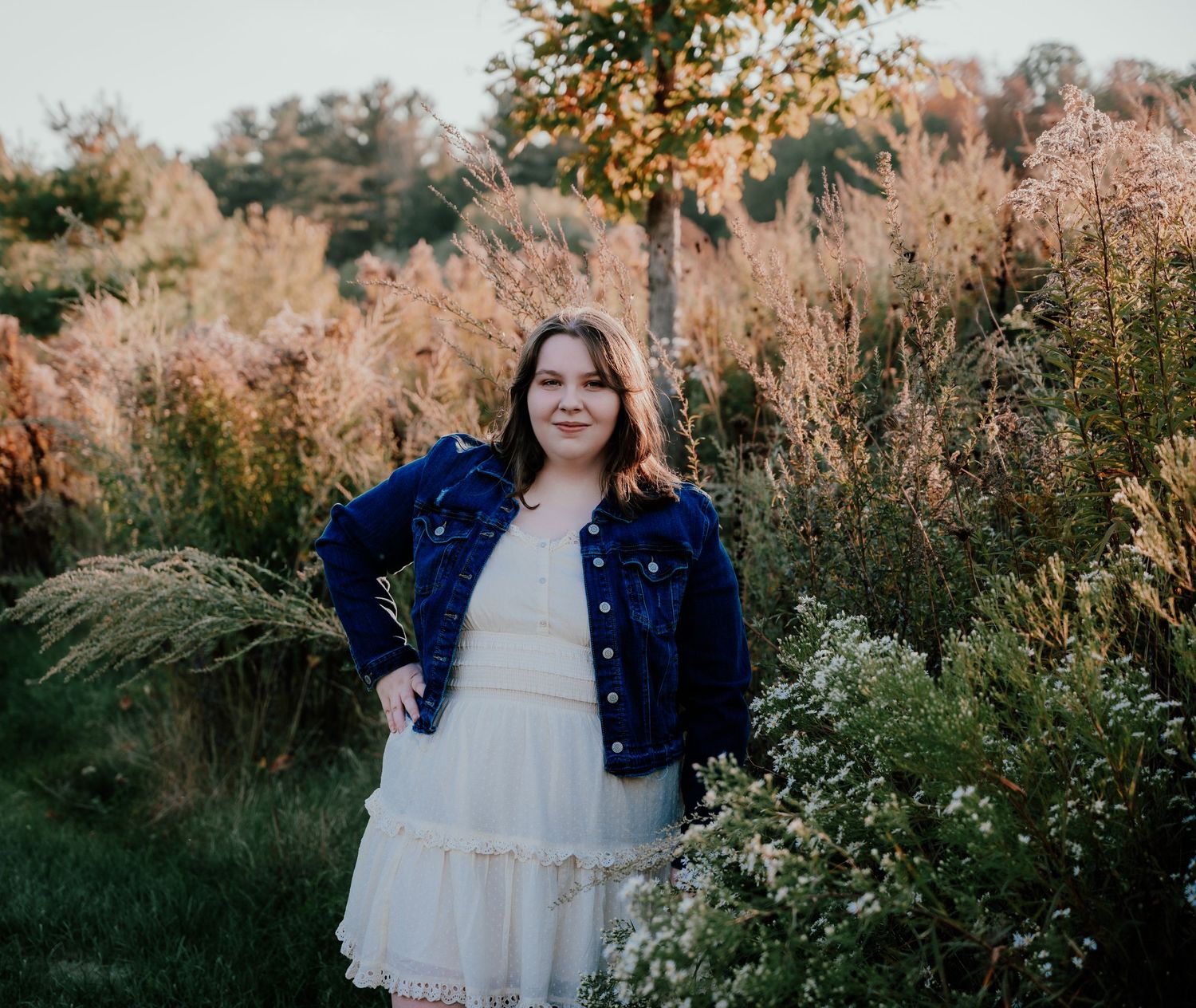 A senior in plaid sits cross-legged on a wooden dock by a serene lake surrounded by autumn trees.