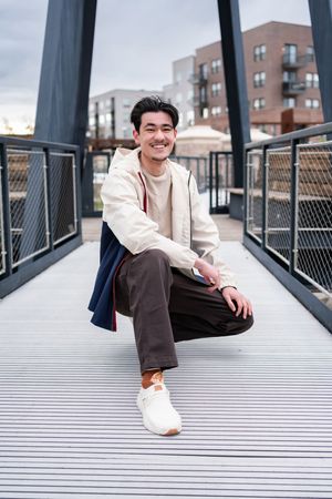 A person in casual attire sits smiling on an urban footbridge wearing white sneakers and layered clothing.