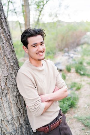 Someone in a beige sweater poses casually outdoors against a tree in a natural woodland setting.