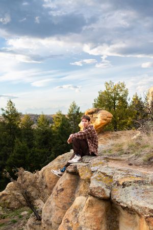 Person sitting on rocky cliff edge overlooking forest landscape under cloudy sky.