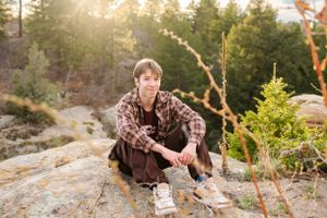 Person in plaid shirt sitting on rocky outcrop during golden hour in mountain forest.