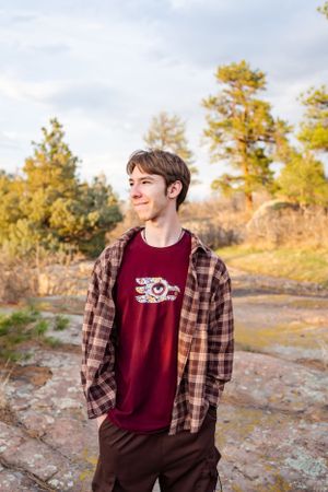 Person in a plaid shirt and burgundy tee stands against a backdrop of autumn trees and rocky terrain.