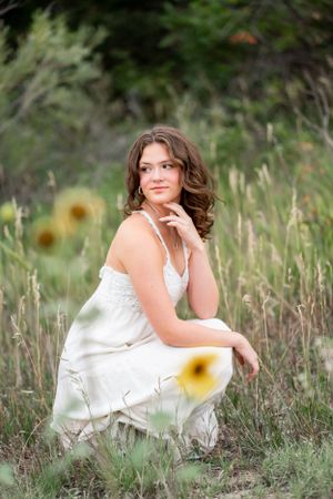 Woman in white dress poses gracefully among tall grass and sunflowers in outdoor setting.