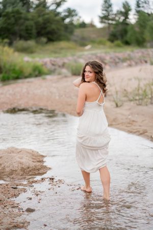 Woman in white dress wades in shallow water along rocky shoreline.