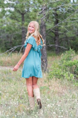 Woman in turquoise dress and boots twirls in a wildflower meadow with pine trees.