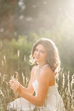 Woman in white dress sits among sunlit grass during golden hour photoshoot.