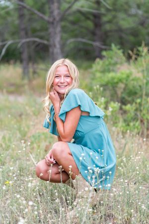 Woman in blue dress crouches among wildflowers in a forest setting.