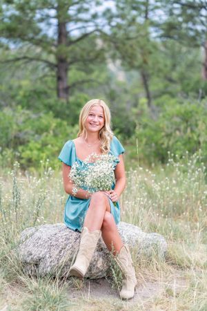Woman in teal dress sits on rock holding wildflowers in mountain setting.