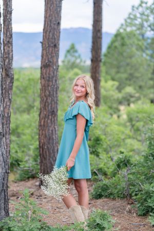 Woman in teal dress holds wildflowers while standing between tall pine trees.