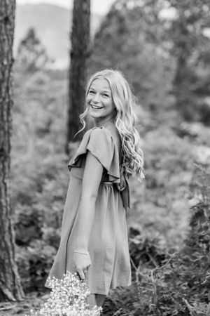 Black and white portrait of woman in dress holding flowers in forest setting.
