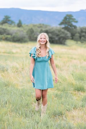 Woman in turquoise dress walks through tall grass with mountains in background.