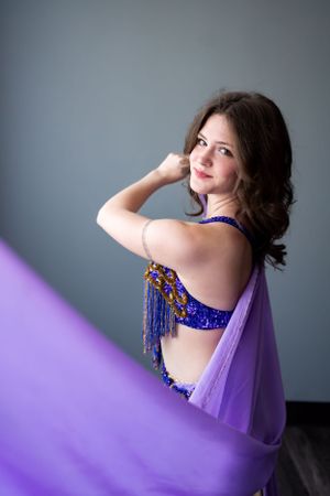 A dance portrait featuring a purple flowing fabric and blue beaded costume against a grey backdrop.