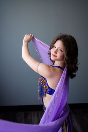 Dancer in purple and blue belly dance costume gracefully poses with flowing veil against gray backdrop.