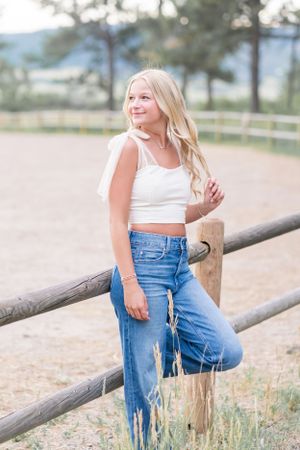 Woman in white crop top and jeans leans against wooden fence in rural setting.