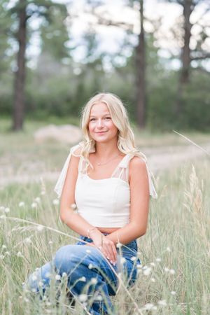 Woman in white top and jeans sits in tall grass with forest backdrop.
