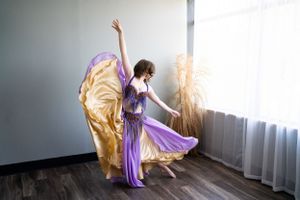 Dancer leaps gracefully in flowing purple and gold costume by sunlit window.