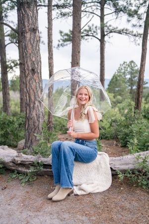 Someone sitting on a fallen log holding a clear umbrella surrounded by tall pine trees.
