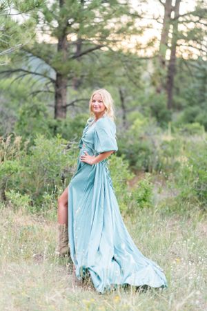 A lighthearted outdoor portrait featuring a flowing blue dress against a pine forest backdrop.