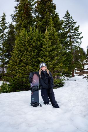A snowboarder in black winter gear poses with their board on a snowy slope surrounded by evergreen trees.