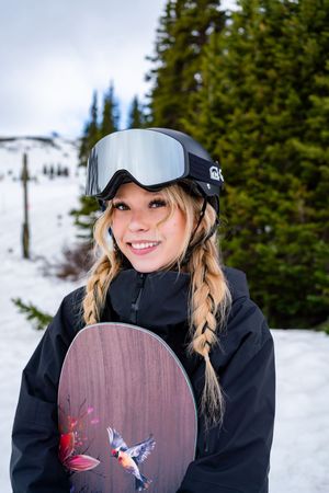 Snowboarder in black jacket and baseball cap holds up board with colorful artwork against snowy mountain backdrop.