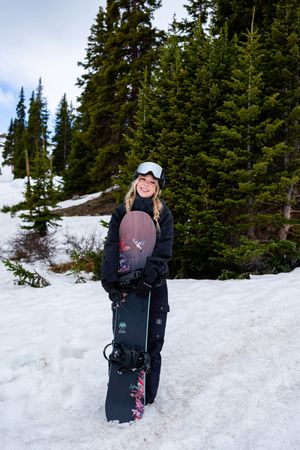 Snowboarder walks through a snowy mountain trail surrounded by evergreen trees on a winter day.
