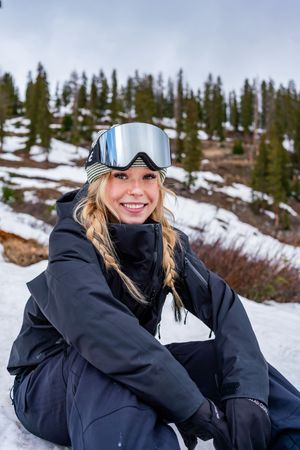Winter sports enthusiast wearing goggles and black snowsuit sits in snowy mountain terrain with evergreen trees behind.