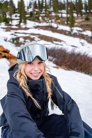 Winter sports enthusiast wearing ski goggles and dark jacket sits in snowy mountain landscape with evergreen trees.