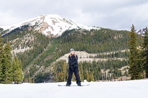 A skier traverses a snowy slope with snow-capped mountains and evergreen forest in the scenic background.