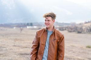 Portrait of someone in a brown leather jacket against a mountainous landscape backdrop.