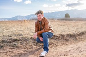 Person in brown leather jacket and jeans sitting on a hillside with mountains in background.