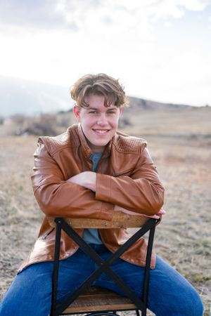 Casual portrait of someone sitting backwards on a chair wearing a brown leather jacket outdoors.