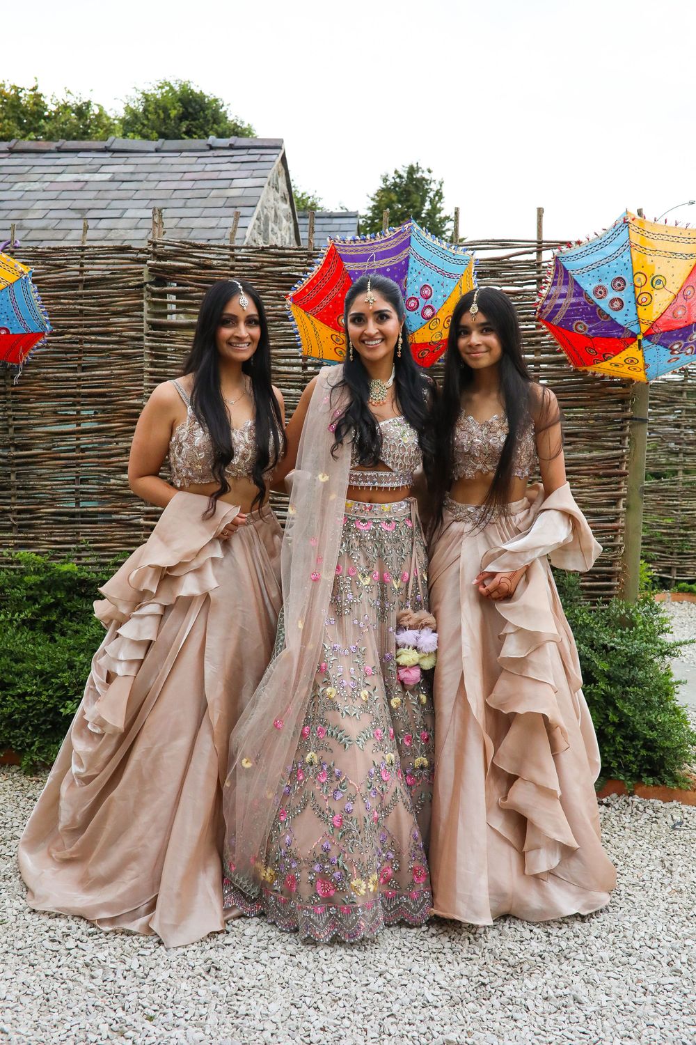 Three women wearing elegant matching beige and floral Indian wedding attire pose with colorful umbrellas in an outdoor setting.